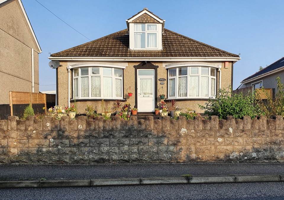 A pebbledash bungalow with a tiled hipped roof behind a low stone wall in Cornwall