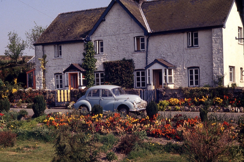 A whitewashed stone cottage with a flower border and a parked car in mid Wales